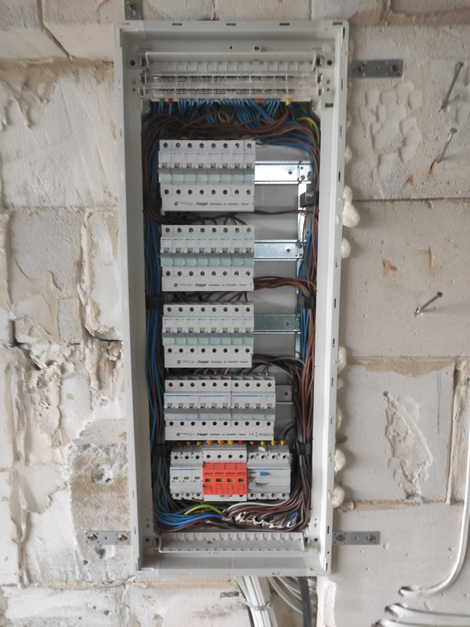Rozdzielnica elektryczna Hager zamontowana na surowej ścianie z widocznymi przewodami, zabezpieczeniami i pianką montażową.