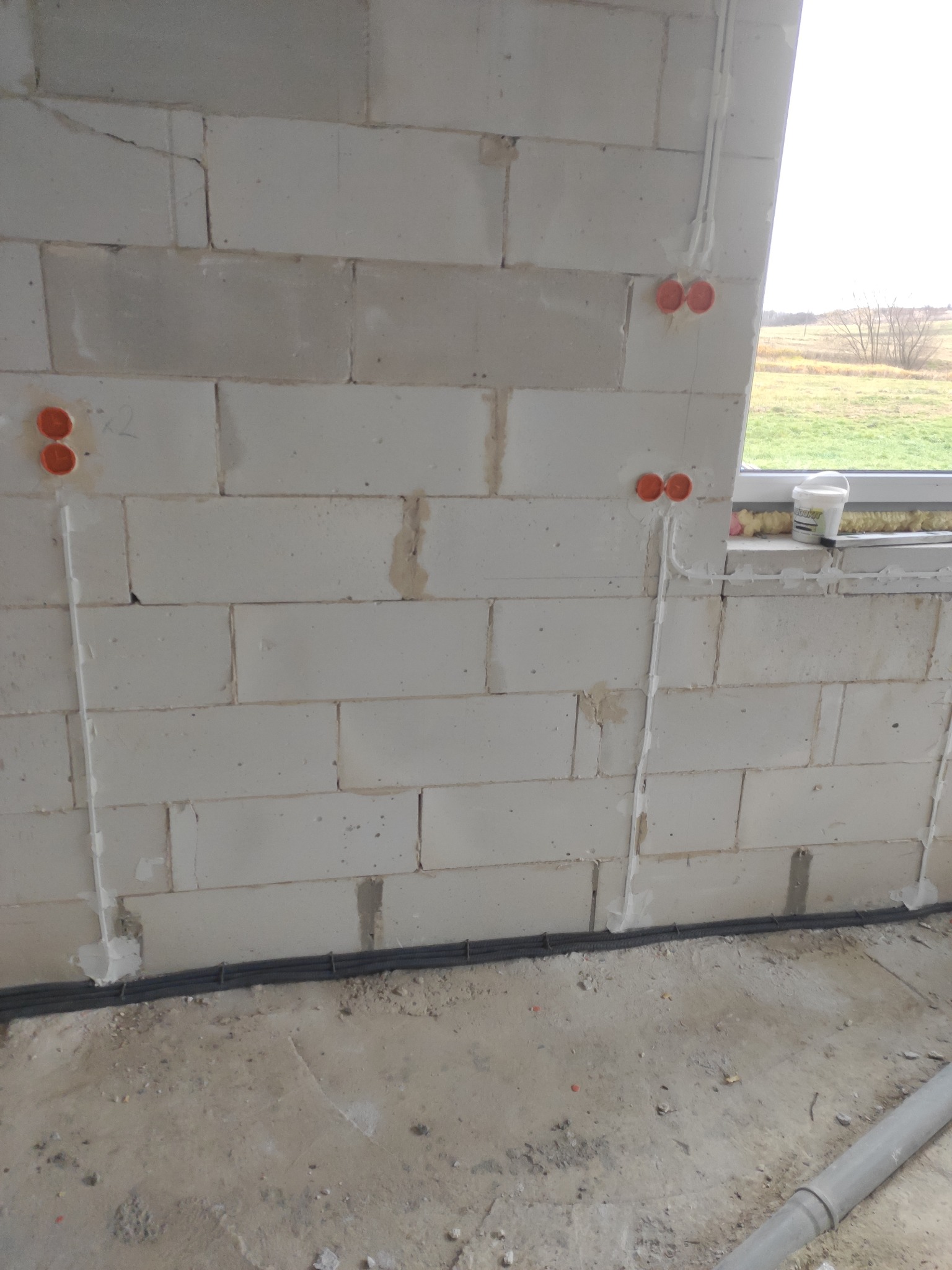 Ściana z betonu komórkowego w trakcie prac instalacyjnych, widoczne peszle i puszki elektryczne, okno w tle.
