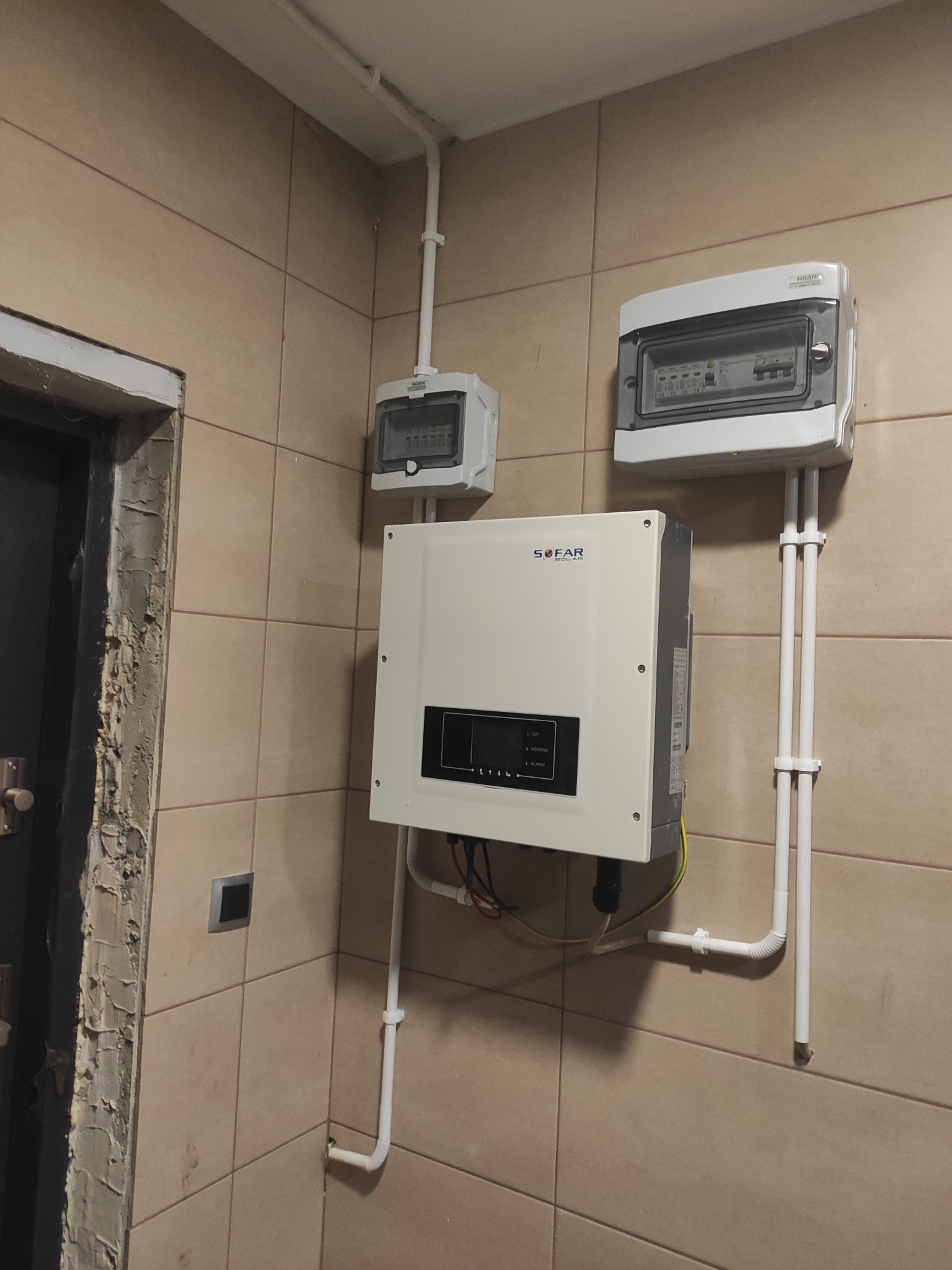 Biały inwerter solarny SOFAR Solar zamontowany na kafelkowanej ścianie, z widocznymi przewodami i skrzynkami elektrycznymi, obok niedokończonego ościeża drzwi.
