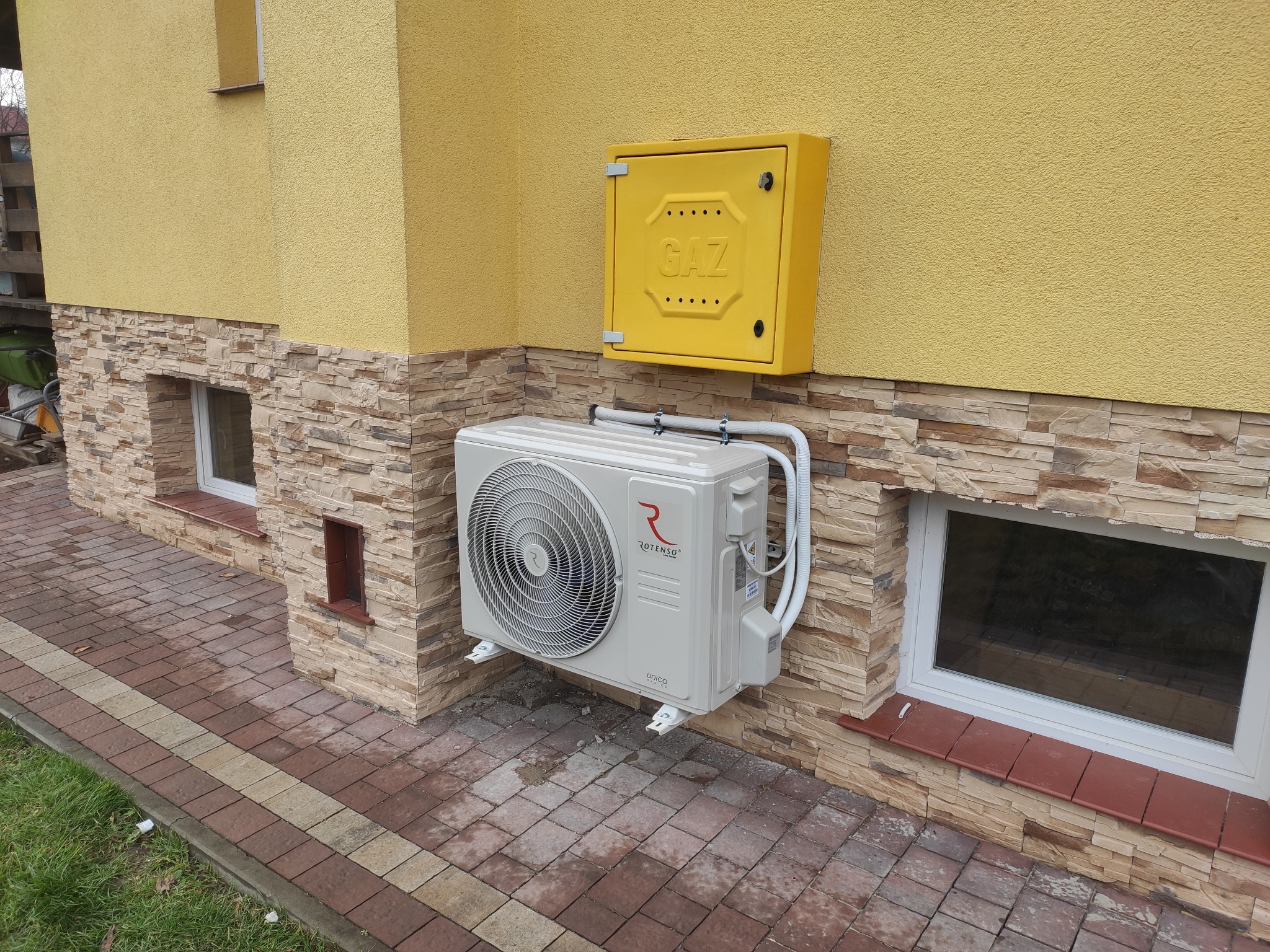 Biała jednostka zewnętrzna klimatyzacji Rotenso Unico zamontowana na elewacji budynku z żółtym tynkiem i okładziną z kamienia, obok żółta skrzynka gazowa.