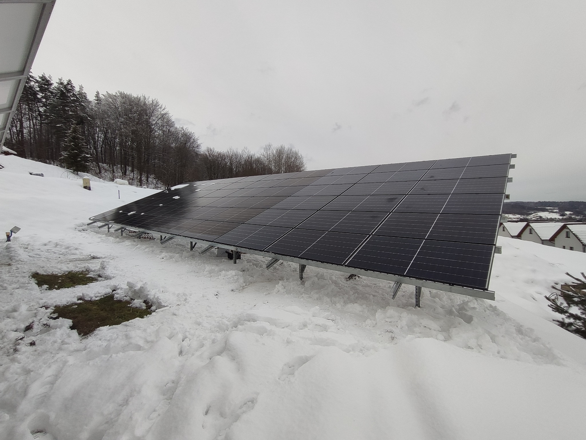 Panele fotowoltaiczne na dachu pokrytym śniegiem, widok z dołu na tle zalesionego wzgórza i zabudowań.