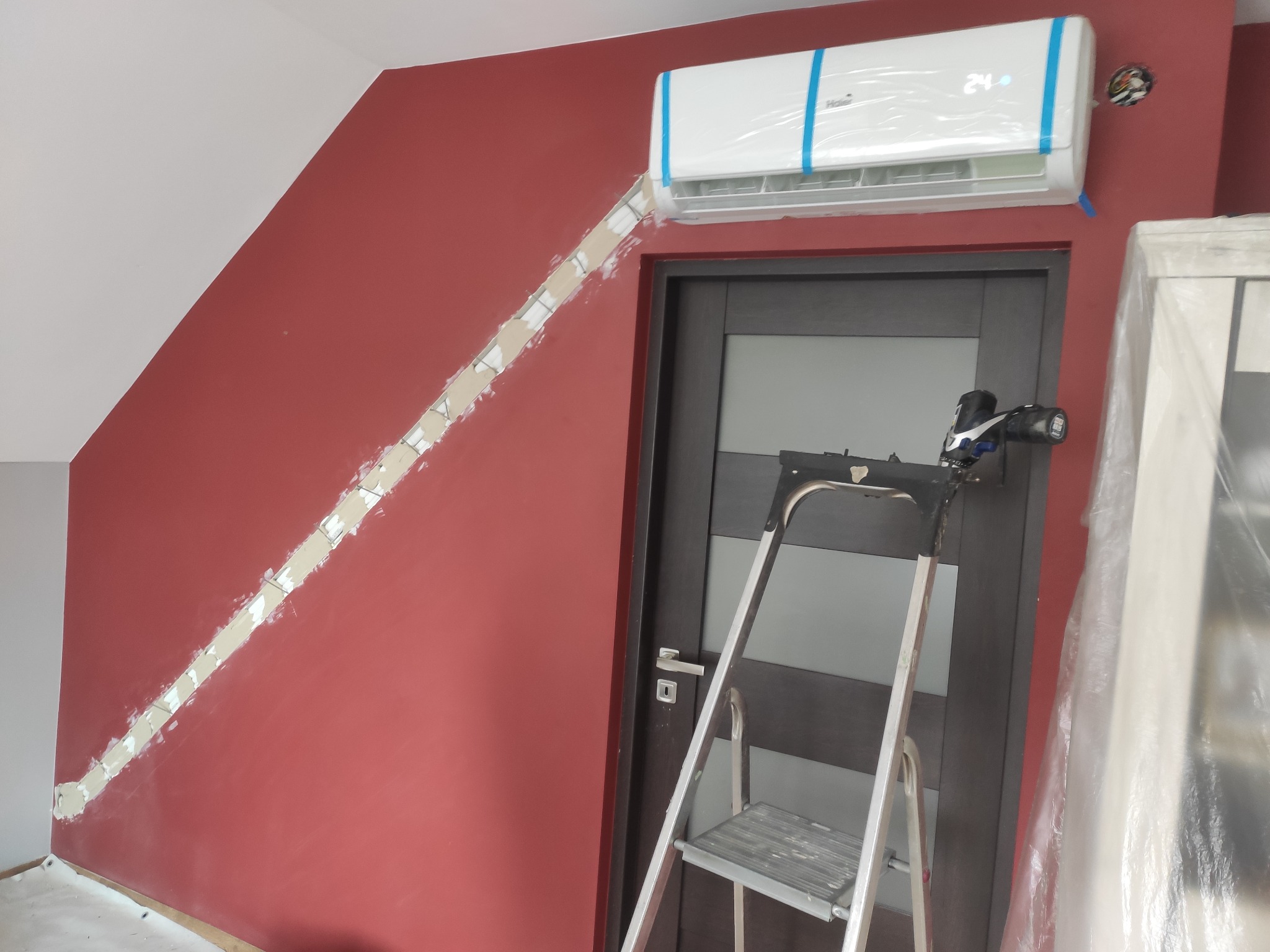 Bordowa ściana z zamontowanym klimatyzatorem Haier w folii ochronnej, widoczna bruzda w ścianie przygotowana do ukrycia instalacji, aluminiowa drabina oparta o ciemne drzwi.