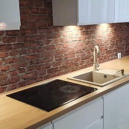 Michał Kulesza - Nowoczesna kuchnia z blatem z jasnego drewna i ceglaną ścianą, oświetlona punktowymi lampami podszafkowymi, z widoczną płytą indukcyjną i beżowym zlewozmywakiem.