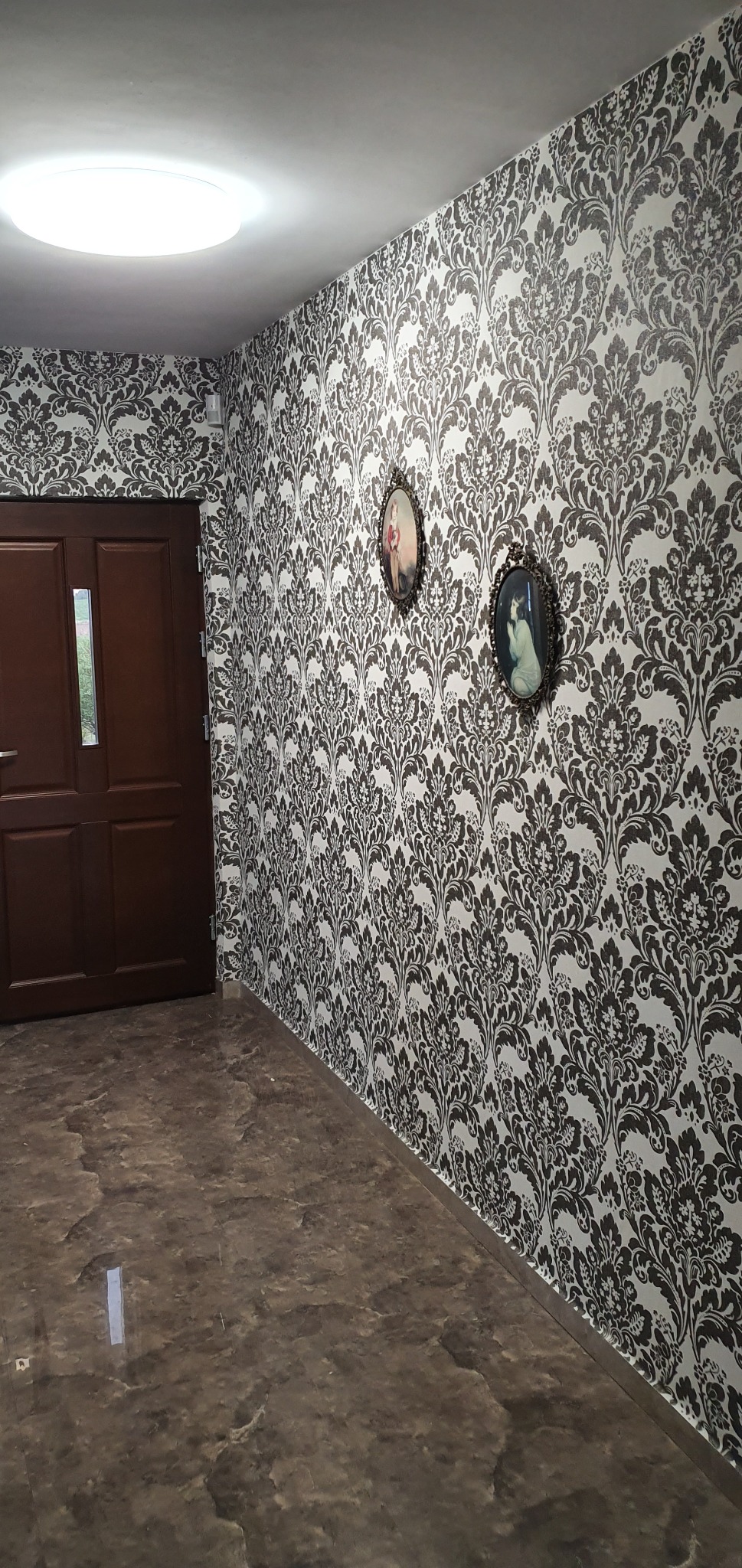 Korytarz z ciemnymi, wzorzystymi tapetami w stylu retro i dwoma owalnymi portretami na ścianie.