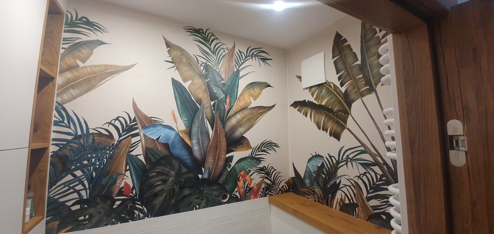 Ściana łazienki z tapetą w motywy tropikalnych liści, widoczny fragment grzejnika drabinkowego i drewnianej ramy drzwi
