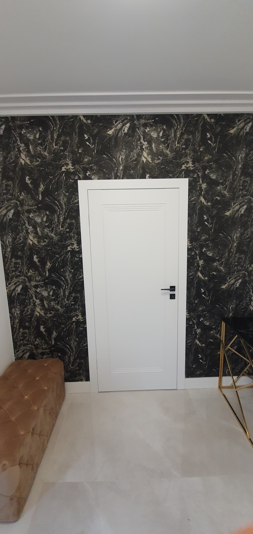 Wnętrze z tapetą imitującą marmur, białe drzwi z czarną klamką, pufa w kolorze musztardowym i stolik z czarnym blatem na złotym stelażu.