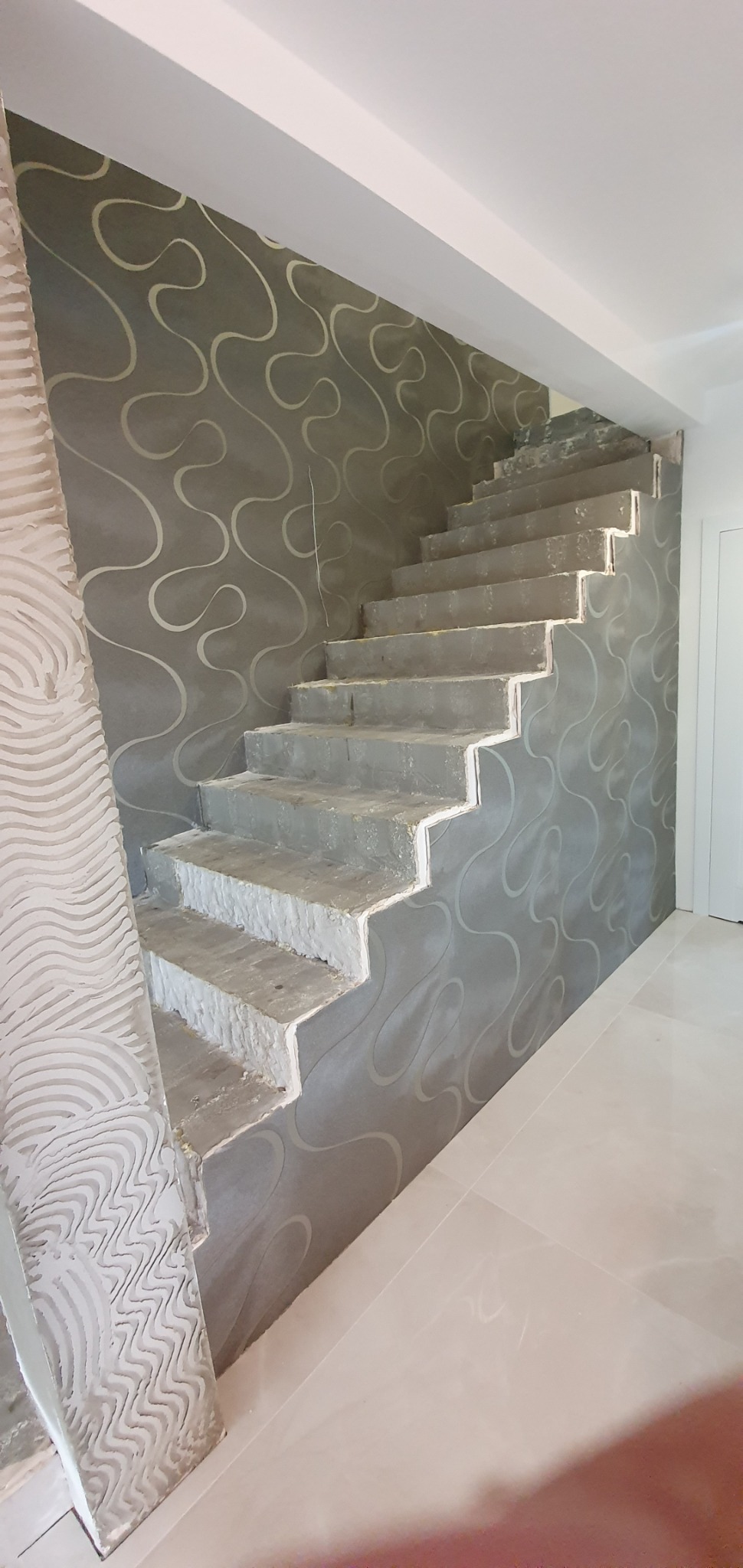Tapetowanie ściany przy schodach w trakcie remontu, widoczny fragment wzorzystej tapety i surowe schody z białymi krawędziami, płytki podłogowe.