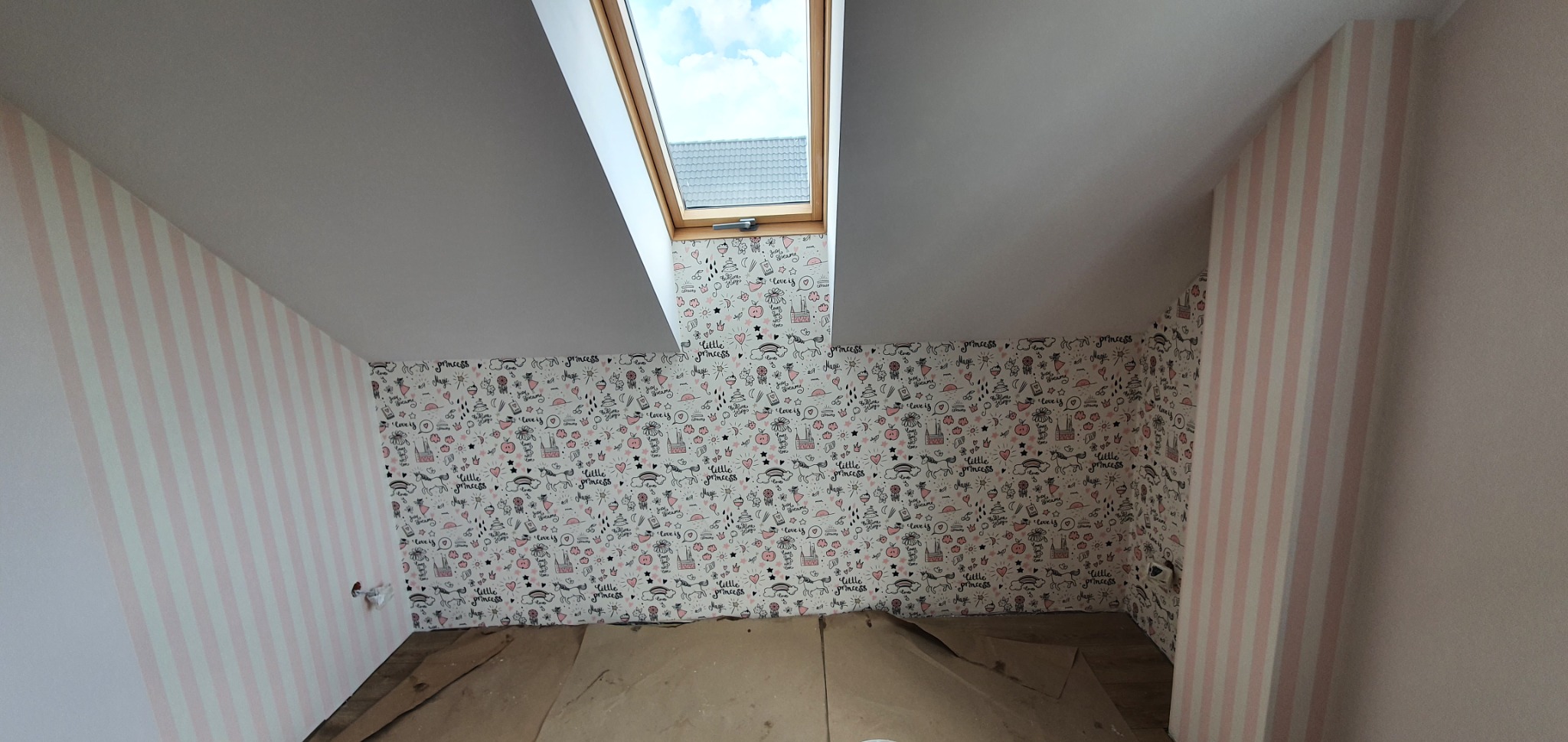 Pokój na poddaszu po częściowym położeniu tapety: biało-różowe pasy na jednej ścianie, wzorzysta tapeta z motywami dziecięcymi na pozostałych, widoczne okno dachowe.