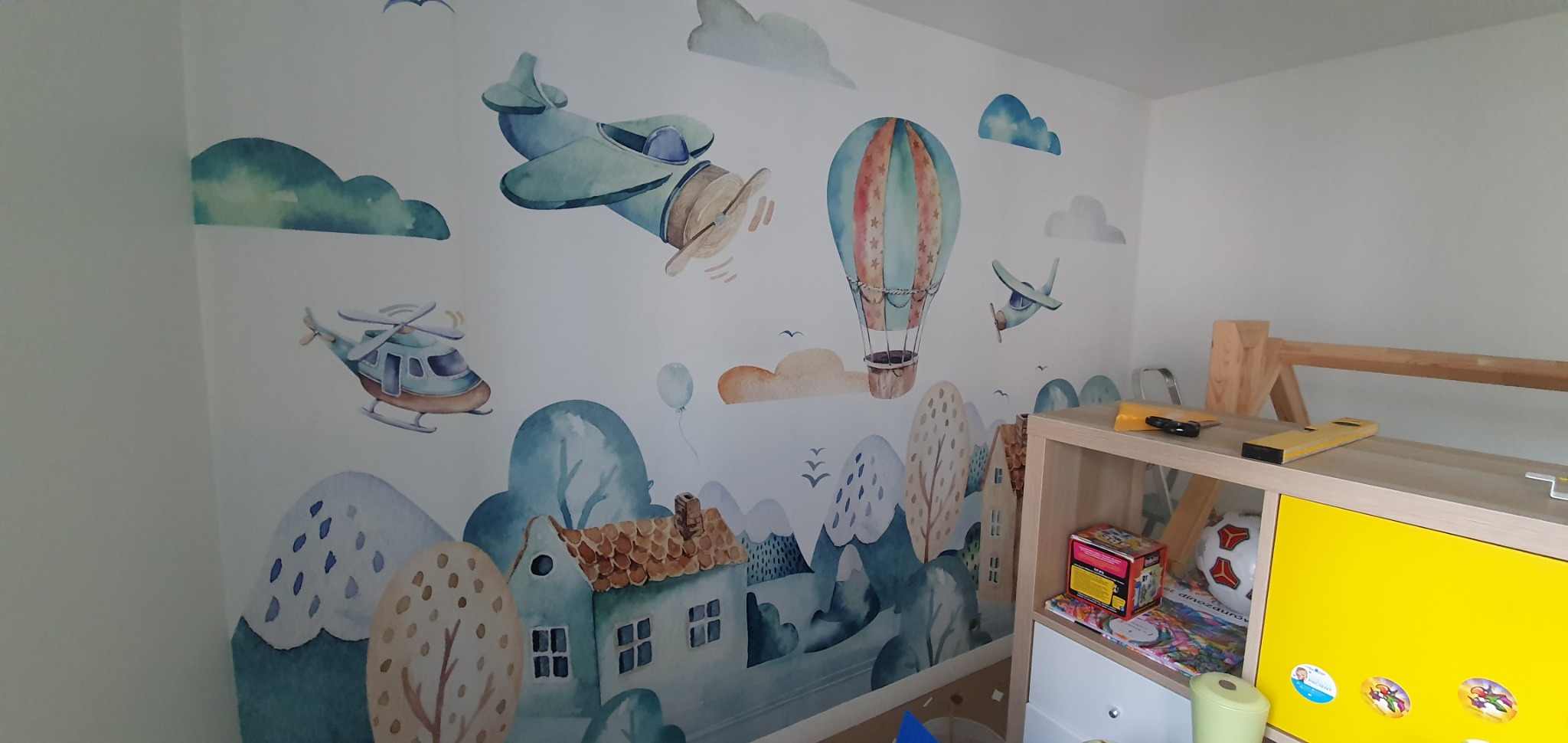 Ściana pokoju dziecięcego z tapetą w pastelowe samoloty, balony i domki, widoczny fragment regału z narzędziami.