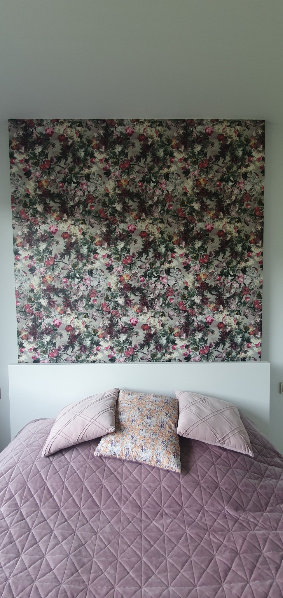 Sypialnia z łóżkiem i ścianą za zagłówkiem pokrytą tapetą w kwiaty, widok z góry na fioletową narzutę i poduszki.