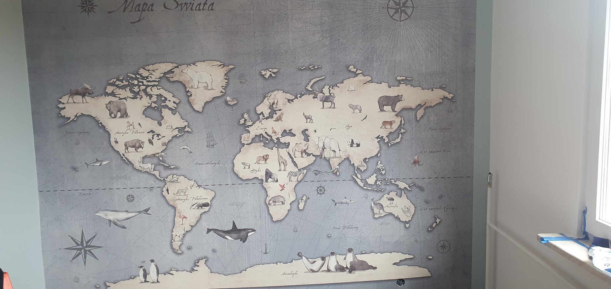 Ściana pokryta szarą tapetą z motywem mapy świata i wizerunkami zwierząt z różnych kontynentów, widoczny fragment ściany i parapetu okiennego.
