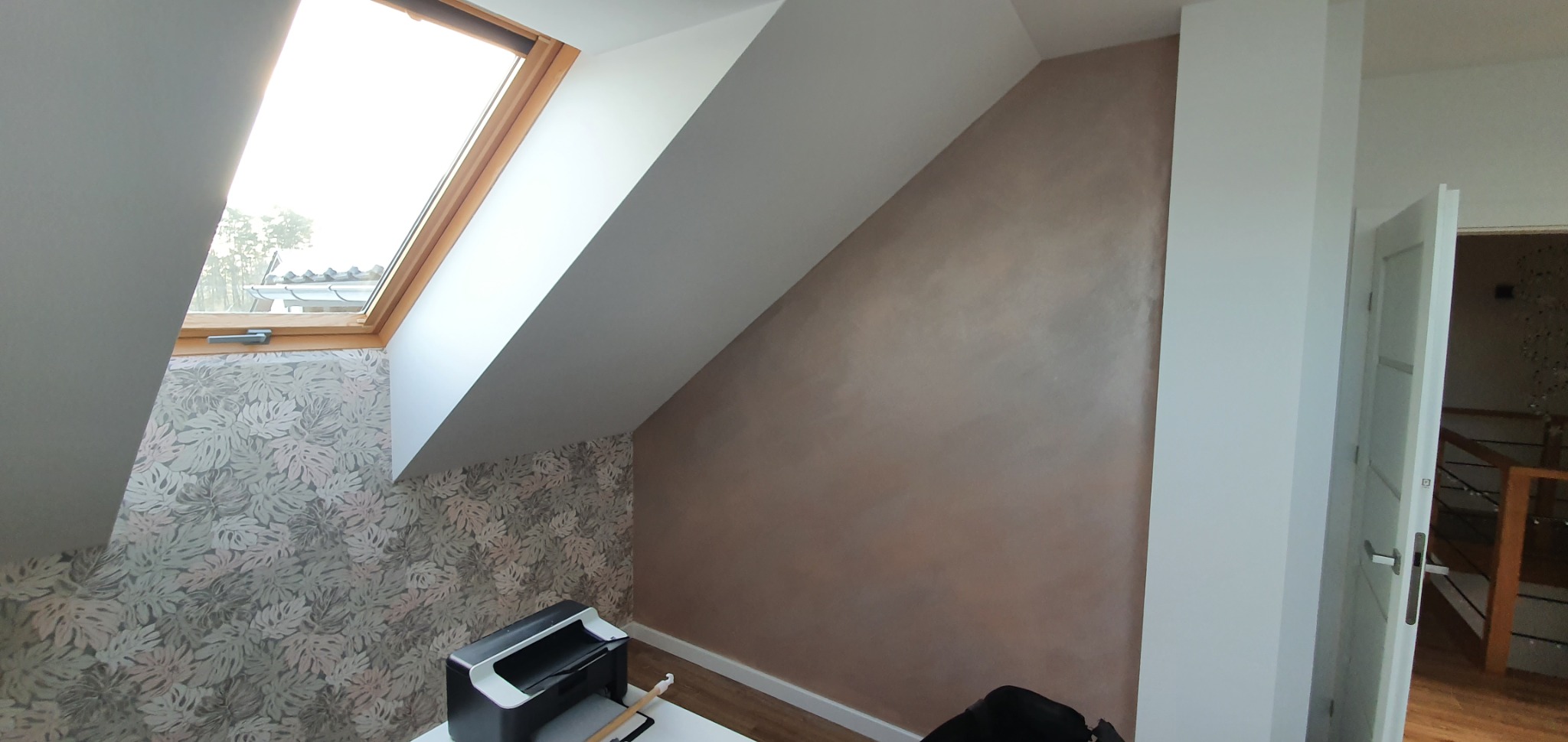 Poddasze po remoncie: ściana z efektem dekoracyjnym w odcieniach brązu, sąsiadująca ze ścianą pokrytą tapetą w liście, okno dachowe w drewnianej ramie.