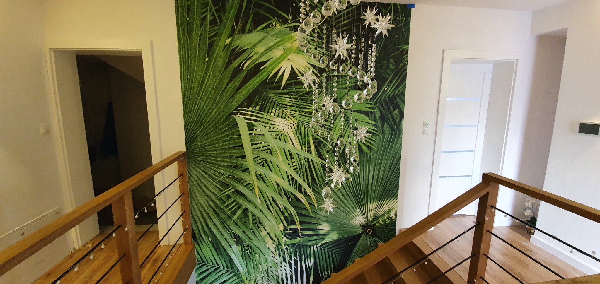 Wnętrze z tapetą w tropikalne liście i kryształowym żyrandolem, widok z drewnianych schodów z balustradą.