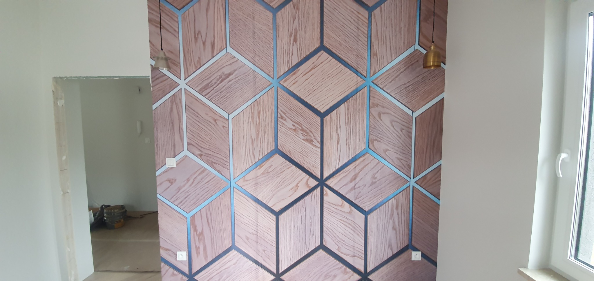 Ściana z tapetą w geometryczny wzór drewnopodobny z niebieskimi akcentami, widoczne wiszące lampy i fragment otwartego przejścia do innego pomieszczenia.