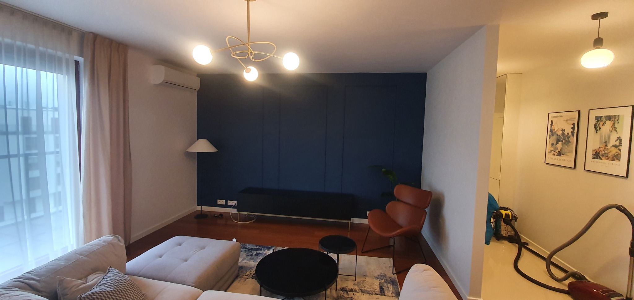 Nowoczesny salon z granatową ścianą z panelami, oświetleniem punktowym i designerską lampą wiszącą; w tle widoczny odkurzacz