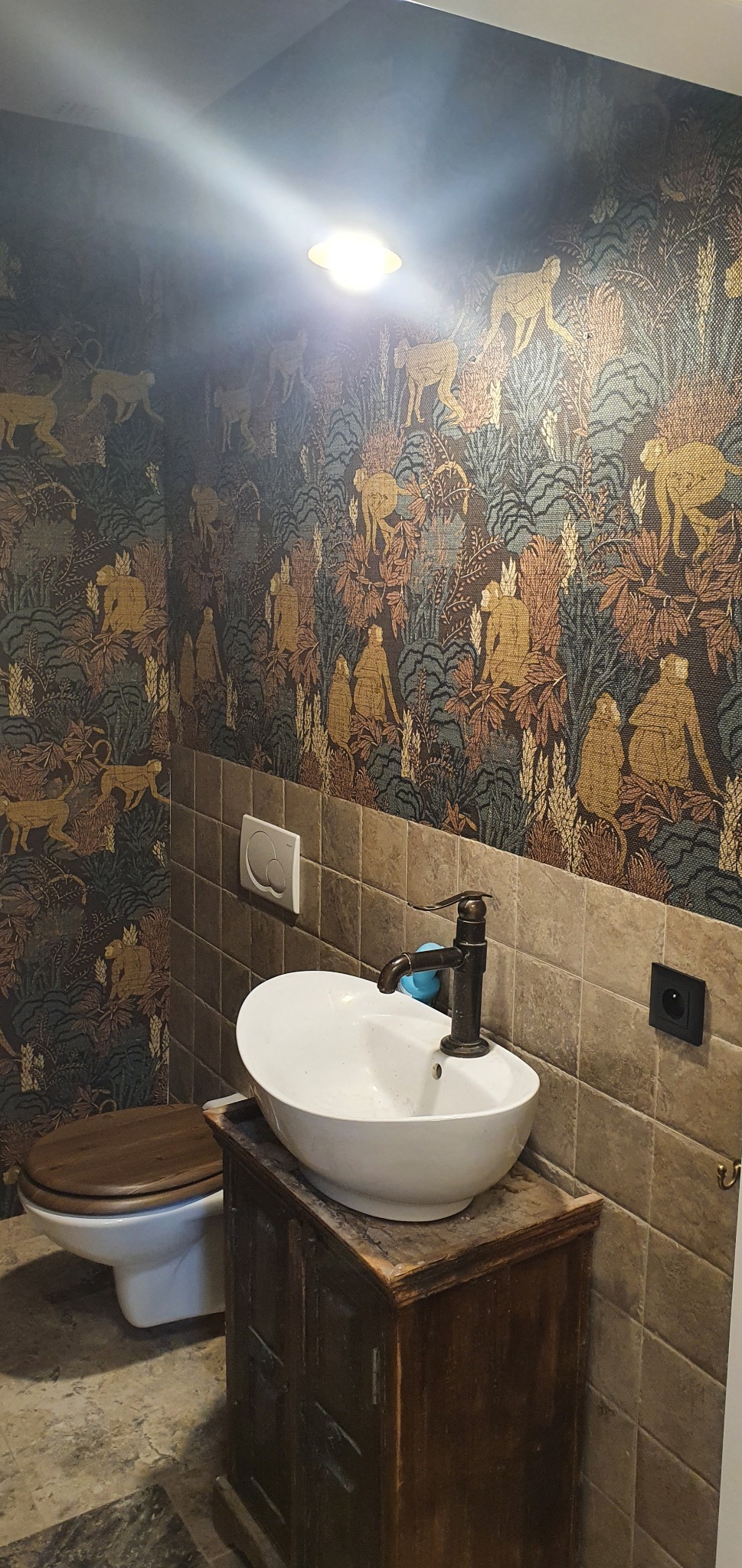 Umywalka nablatowa na postarzanej szafce w łazience w Gdańsku, ściany pokryte wzorzystą tapetą z motywem małp i roślin, widoczna toaleta z drewnianą deską.