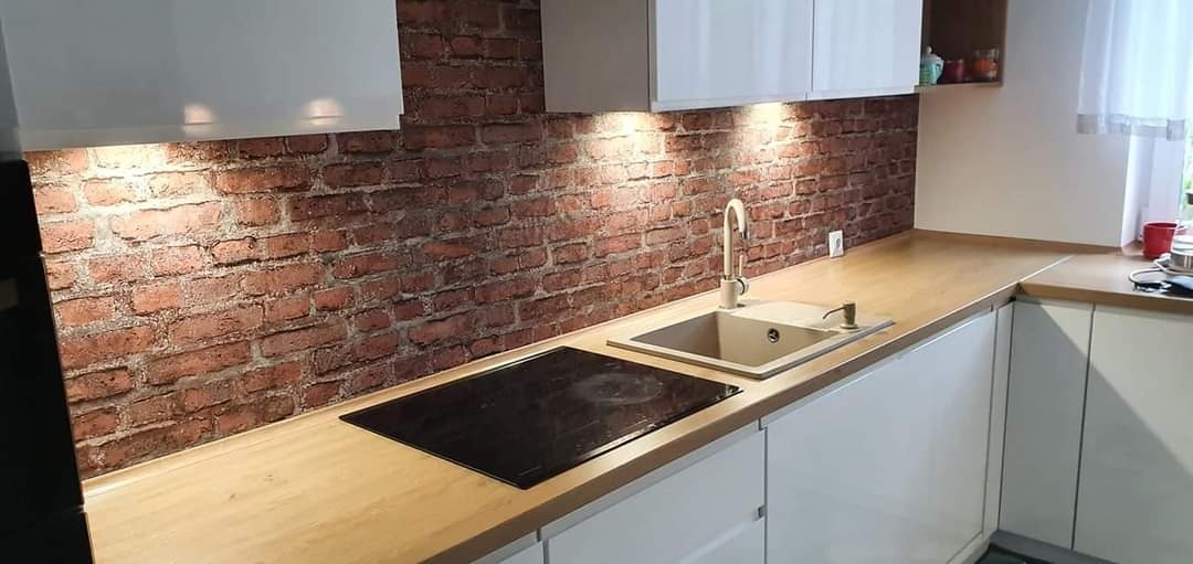 Nowoczesna kuchnia z blatem z jasnego drewna i ceglaną ścianą, oświetlona punktowymi lampami podszafkowymi, z widoczną płytą indukcyjną i beżowym zlewozmywakiem.