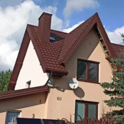 Dom jednorodzinny z brązowym dachem z dachówki ceramicznej, kominem i widocznym fragmentem konstrukcji więźby dachowej po demontażu poszycia, na tle błękitnego nieba z chmurami.