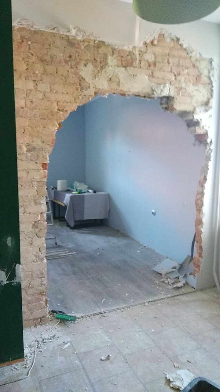 Otwór w ścianie z cegły, widok na pokój z niebieskimi ścianami i stolikiem przykrytym szarą tkaniną, podłoga częściowo zakurzona gruzem, etap prac remontowych.