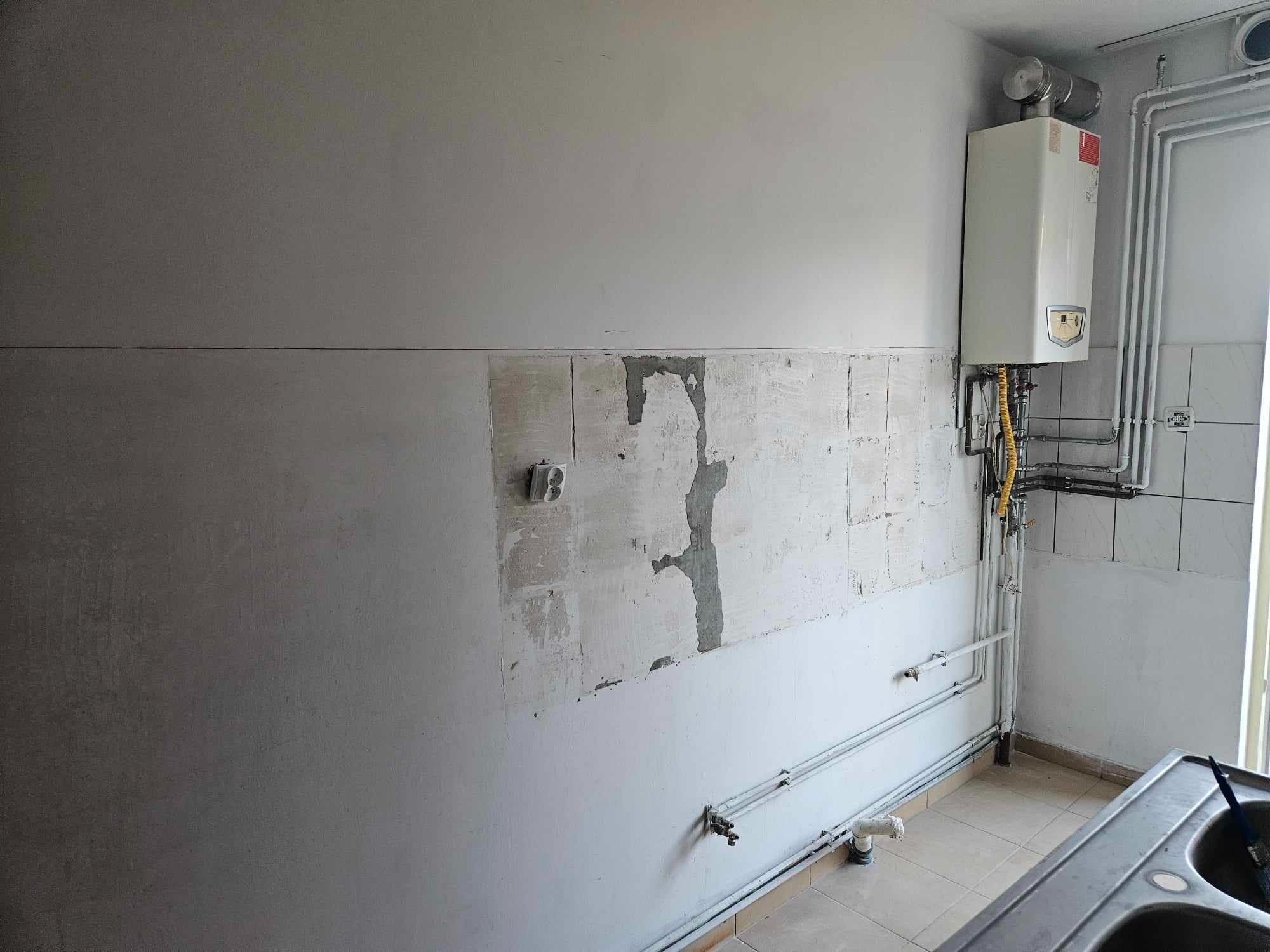 Ściana w trakcie przygotowania do położenia płytek w łazience, widoczne ślady po wcześniejszych płytkach, gładka ściana powyżej i wiszący piec gazowy.