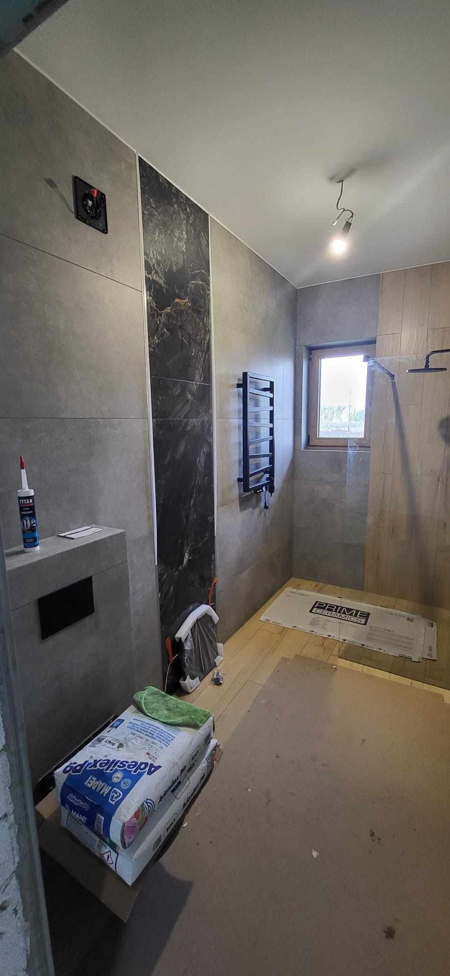 Wnętrze łazienki w trakcie prac wykończeniowych: położone szare płytki, panel dekoracyjny imitujący marmur, grzejnik drabinkowy, przygotowane worki z klejem do płytek MAPEI Adesilex P9 oraz narzędzia.