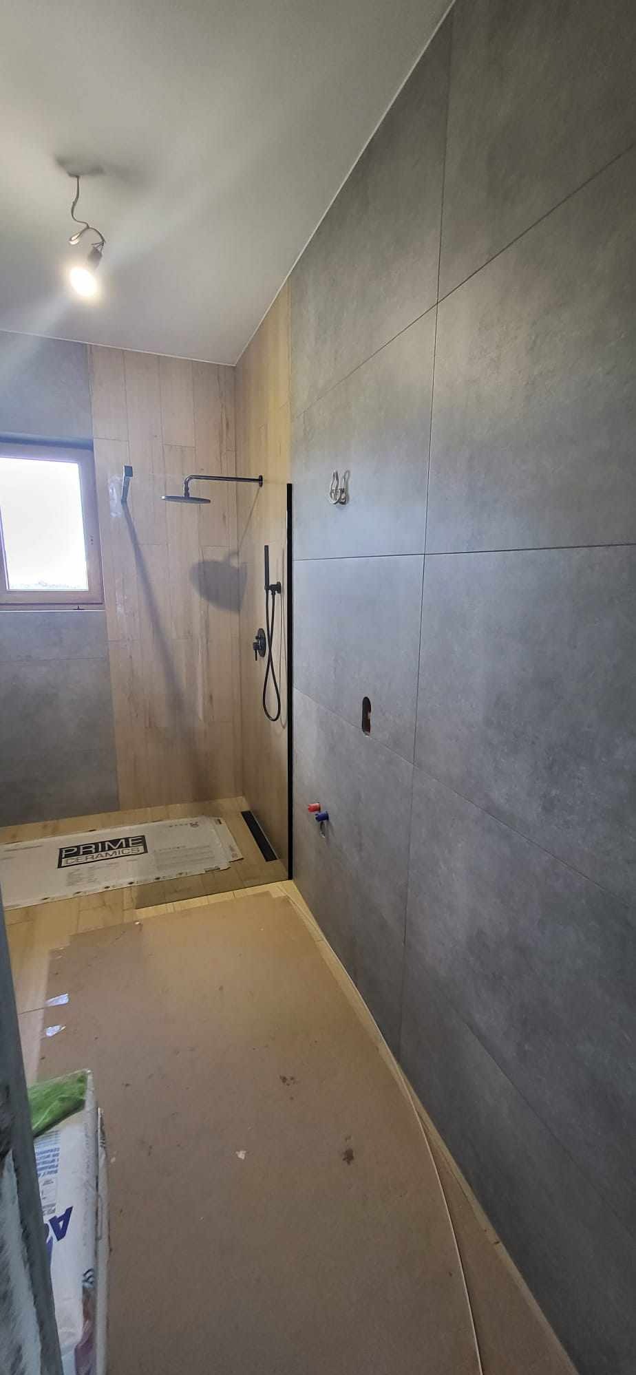 Nowoczesna łazienka w trakcie wykańczania: szare, wielkoformatowe płytki na ścianie, drewnopodobne płytki w strefie prysznica, widoczne instalacje wodne i odpływ liniowy. Podłoga zabezpieczona...