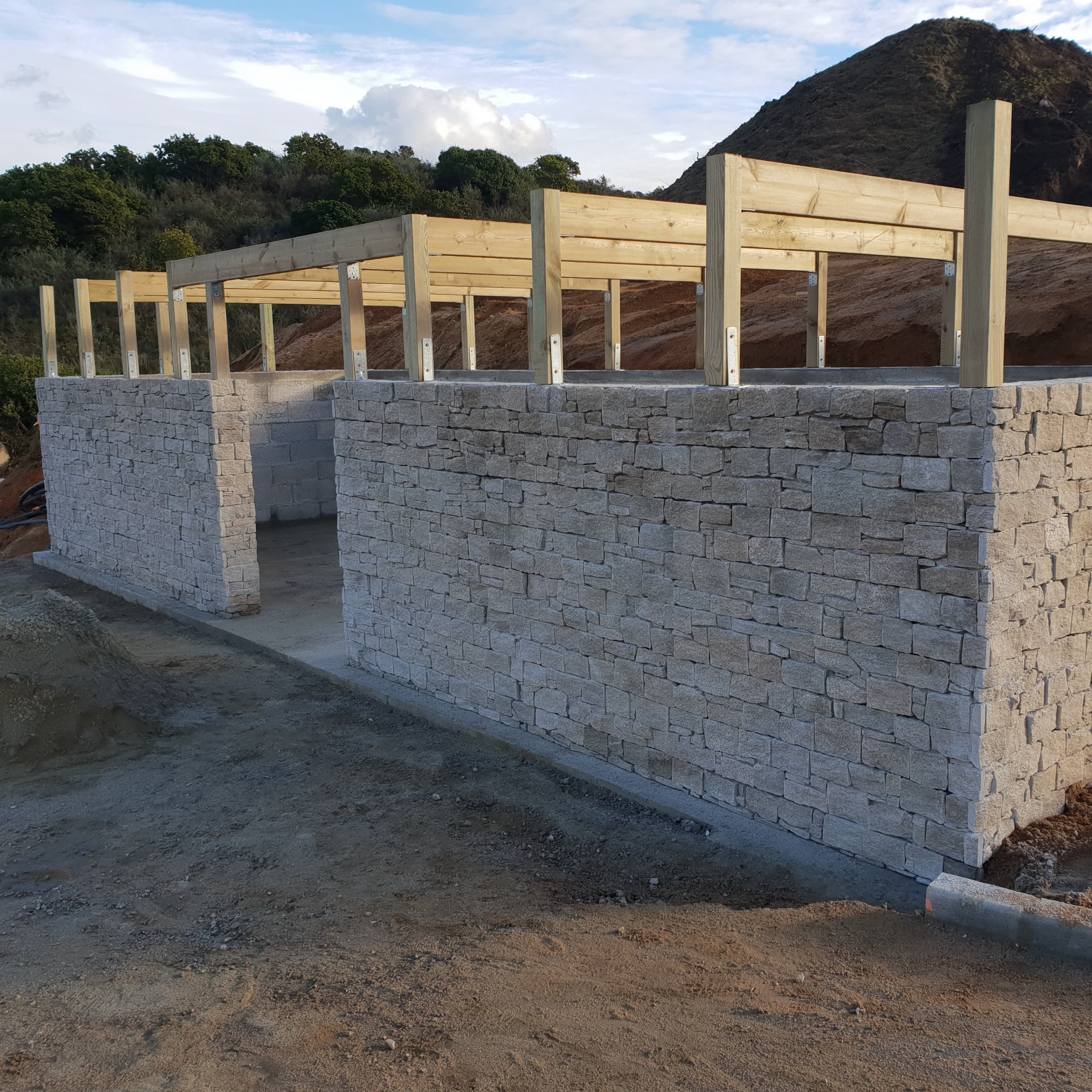 Surowe ściany z kamienia łupanego w trakcie budowy, z widoczną drewnianą konstrukcją dachu i podłożem z piasku, na tle pagórkowatego krajobrazu.