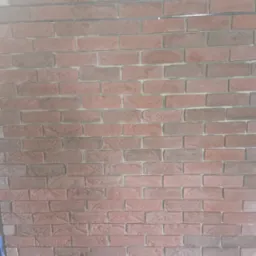 Ściana wyłożona cegłą klinkierową w odcieniach czerwieni i brązu, widoczne fragmenty instalacji elektrycznej i niebieskiej folii zabezpieczającej.