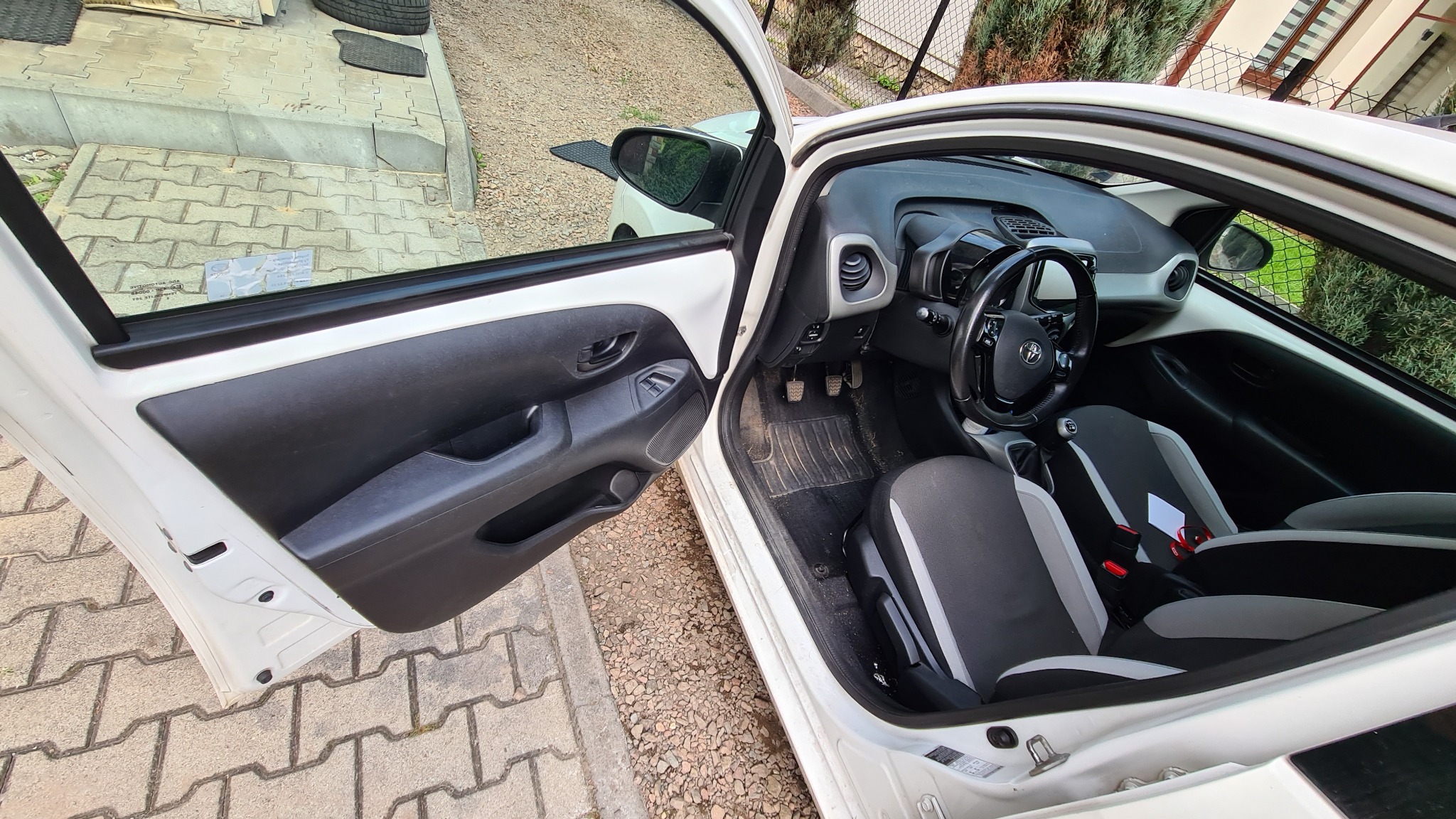 Wnętrze białego samochodu z otwartymi drzwiami, widoczne szaro-czarne fotele i brudna wycieraczka pod nogami kierowcy.