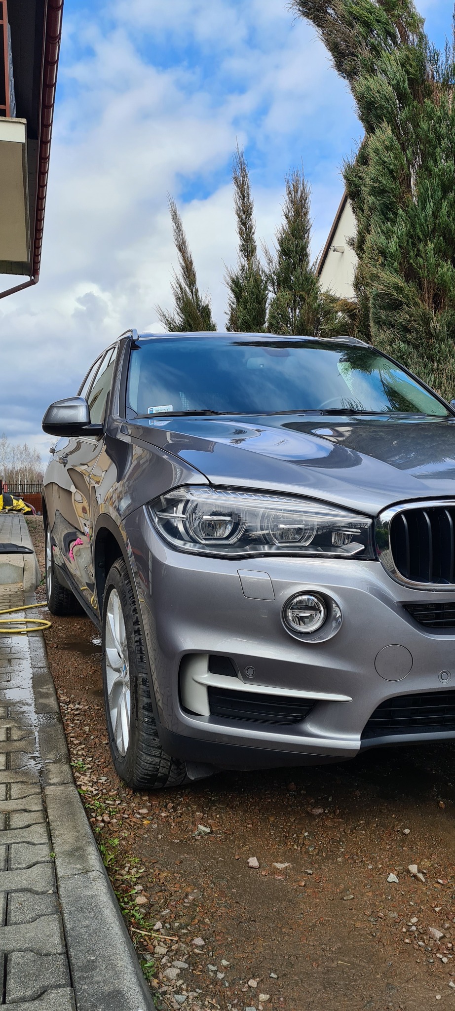 Szary SUV marki BMW, model X5, po umyciu, z widoczną żółtą rurą od myjki ciśnieniowej leżącą na mokrym chodniku.