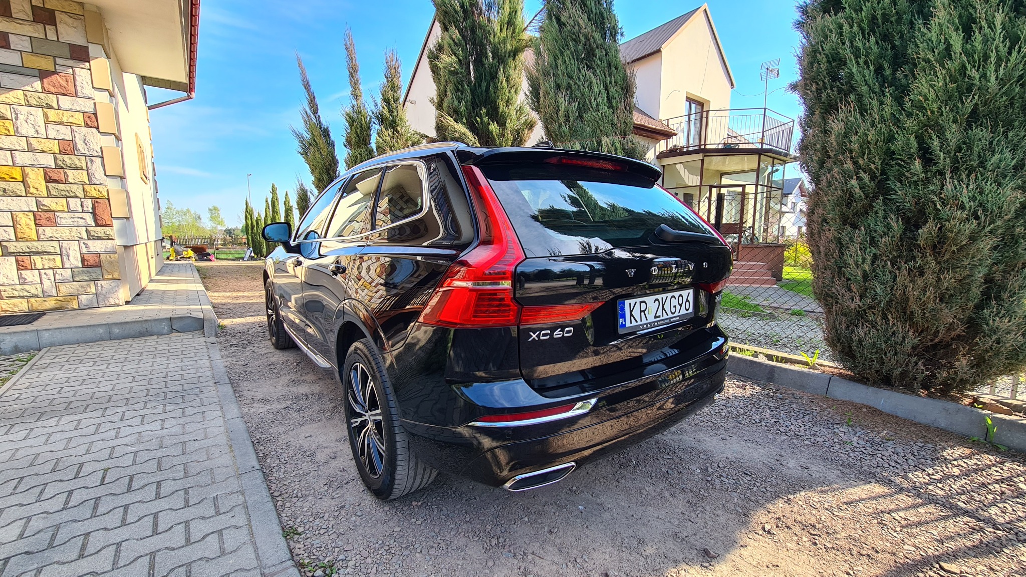 Błyszczący, czarny samochód Volvo XC60 zaparkowany na żwirowym podjeździe przed domem z ceglaną elewacją, widok z tyłu z numerem rejestracyjnym KR 2KG96