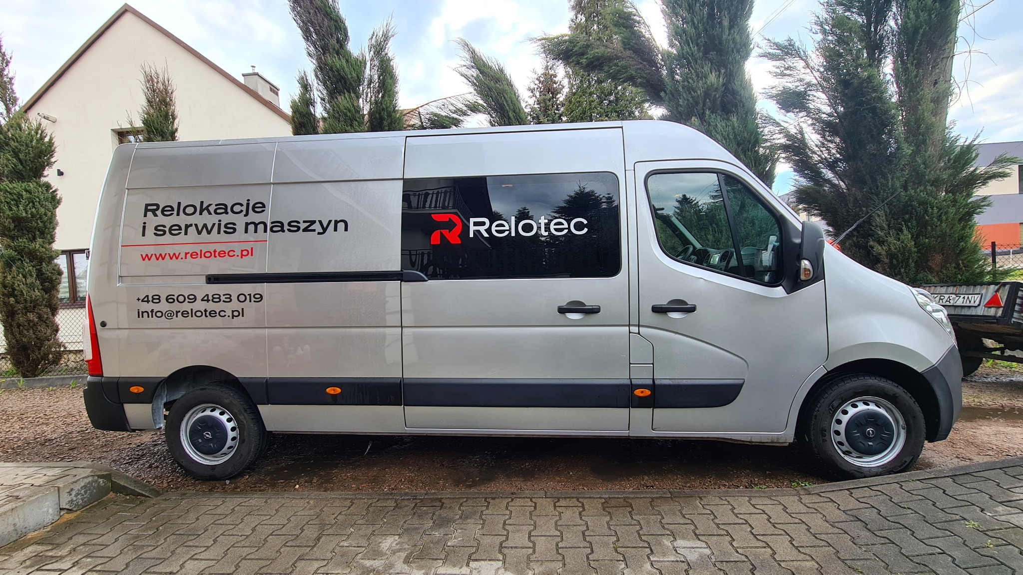 Srebrny samochód dostawczy z napisem 'Relokacje i serwis maszyn' oraz logiem firmy 'Relotec' na tle budynku i drzew.