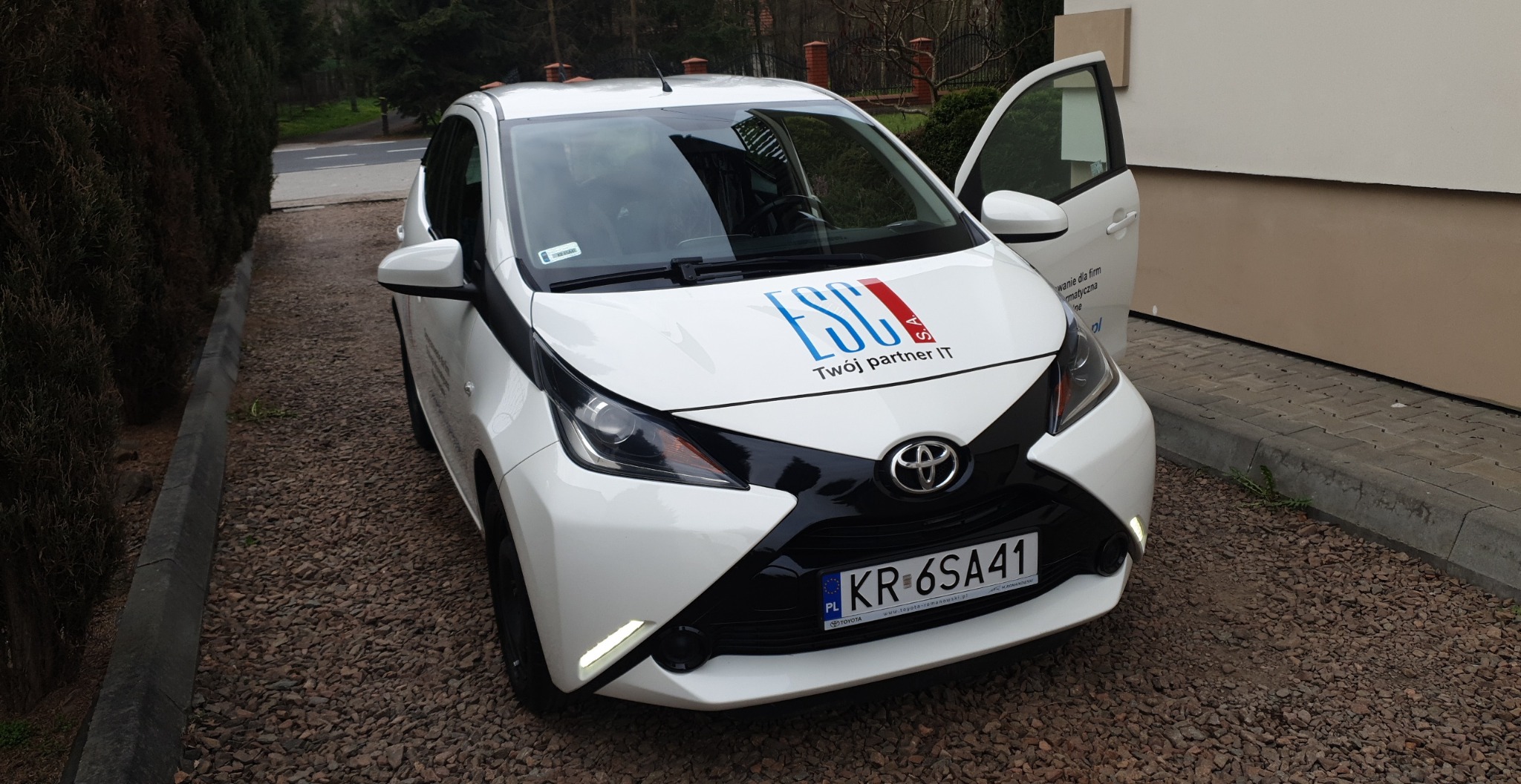 Biały samochód osobowy Toyota z logo firmy ESC S.A. na masce, zaparkowany na żwirowym podjeździe, z otwartymi drzwiami pasażera.