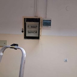 Oscar Kamil Osinski - Otwarta skrzynka z bezpiecznikami na ścianie w dwukolorowym pomieszczeniu, widoczny fragment drabiny aluminiowej z czarnym uchwytem na górze, obok skrzynka z napisem 'PACZKOMAT'.