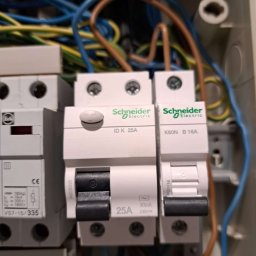 Oscar Kamil Osinski - Szczegółowe ujęcie tablicy rozdzielczej z bezpiecznikami Schneider Electric, widoczne przewody w kolorach niebieskim, brązowym i żółto-zielonym, czarne przełączniki w pozycji wyłączonej.