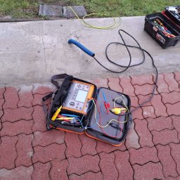 Oscar Kamil Osinski - Otwarta torba narzędziowa z miernikiem elektrycznym i przewodami pomiarowymi leżąca na czerwonej kostce brukowej, obok skrzynka narzędziowa i żółty przewód na betonowym chodniku, trawa w tle.