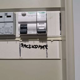 Oscar Kamil Osinski - Tablica rozdzielcza z wyłącznikami, opisana markerem słowem 'PACZKOMAT' na białej obudowie.