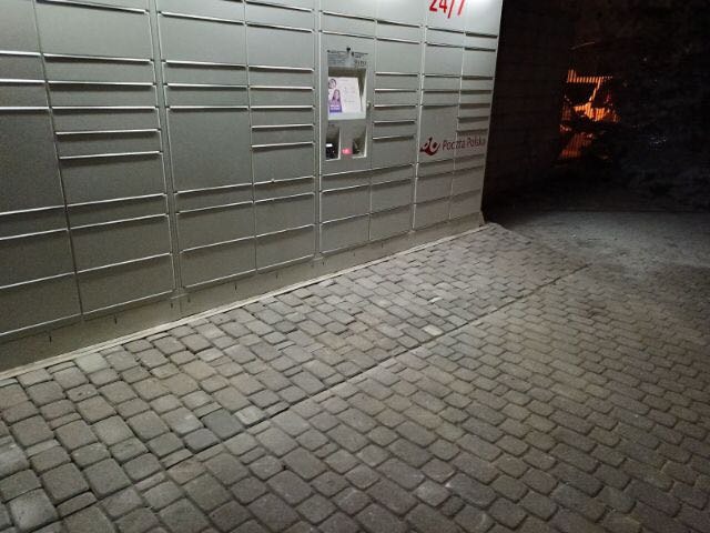 Szara paczkomat Poczty Polskiej na tle brukowanej nawierzchni, widok nocny.