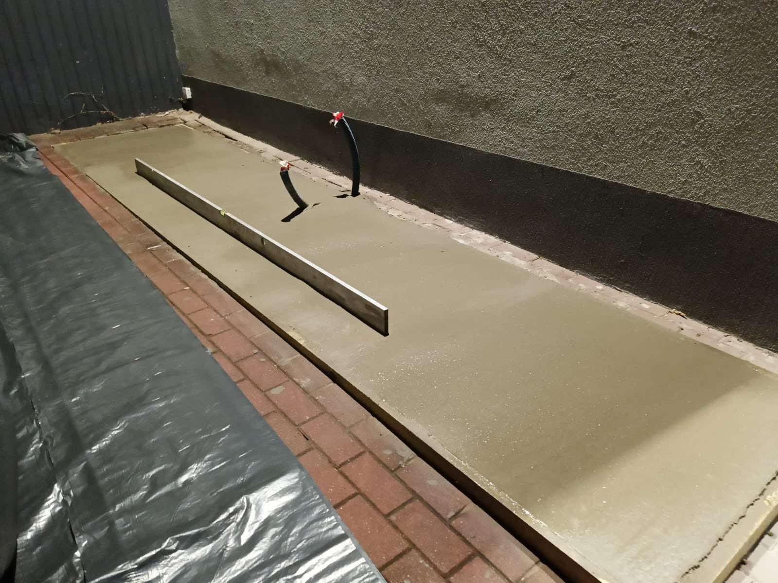 Świeżo wylana, wyrównana wylewka betonowa na zewnątrz budynku, z wystającymi rurami instalacji i aluminiową łatą poziomującą, częściowo przykryta folią ochronną.