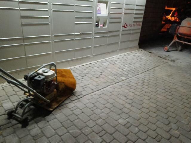 Zagęszczarka spalinowa na nowo ułożonej kostce brukowej przed paczkomatem Poczty Polskiej, widoczna betoniarka w tle, wieczorne oświetlenie.