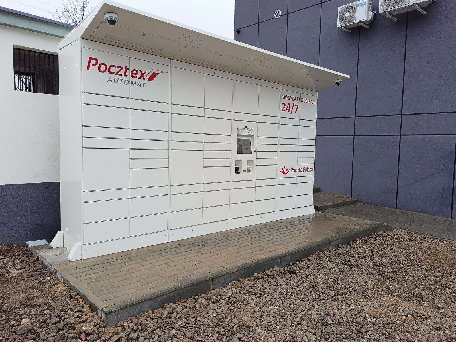 Automatyczna skrzynka pocztowa Pocztex z napisem 'Wysyłaj i odbieraj 24/7' na tle nowoczesnego budynku z szarą elewacją.
