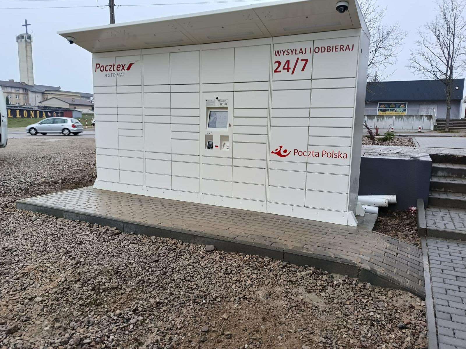Biały automat pocztowy Pocztex z ekranem dotykowym i logo Poczty Polskiej, umieszczony na zewnątrz budynku z kostki brukowej i żwiru.