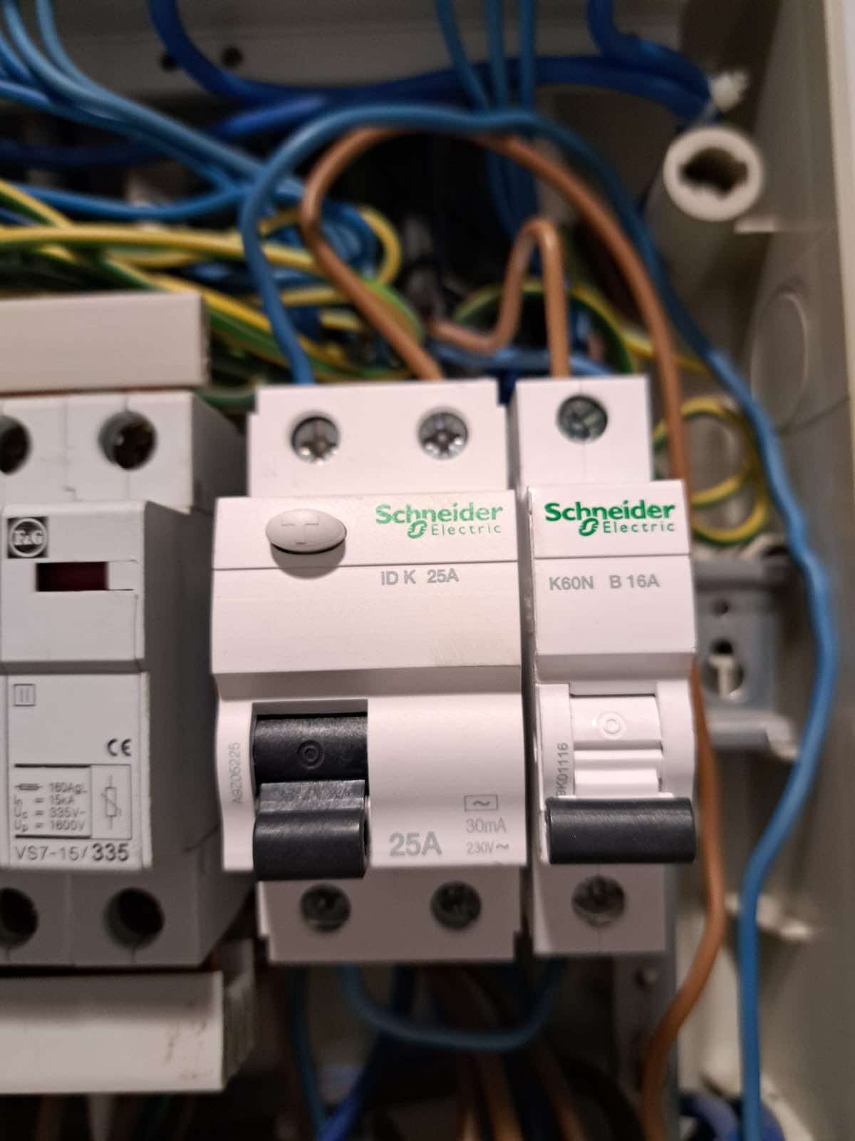 Szczegółowe ujęcie tablicy rozdzielczej z bezpiecznikami Schneider Electric, widoczne przewody w kolorach niebieskim, brązowym i żółto-zielonym, czarne przełączniki w pozycji wyłączonej.