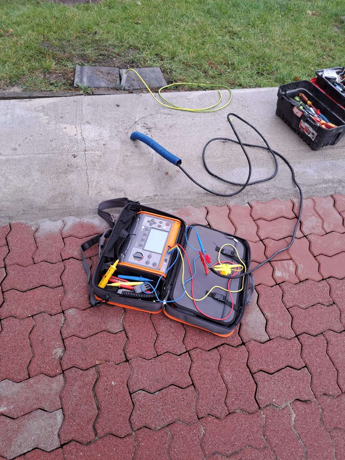 Otwarta torba narzędziowa z miernikiem elektrycznym i przewodami pomiarowymi leżąca na czerwonej kostce brukowej, obok skrzynka narzędziowa i żółty przewód na betonowym chodniku, trawa w tle.