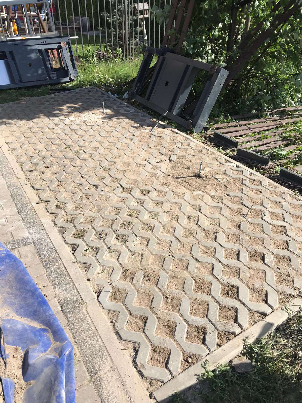 Ułożona, betonowa ażurowa kostka brukowa z wypełnieniem ziemią i fragmentami trawy, widoczne elementy obudowy studzienki kanalizacyjnej i zarośla w tle.