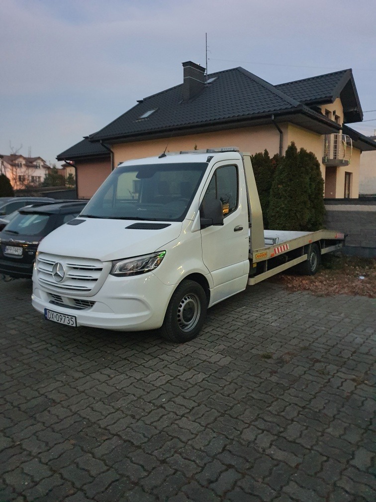 Biała laweta Mercedes-Benz Sprinter zaparkowana na brukowanym podjeździe przed domem z ciemną dachówką, widoczny numer rejestracyjny.