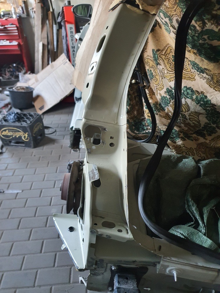 Fragment nadwozia samochodu w trakcie naprawy blacharskiej, widoczne spawy i elementy karoserii w jasnym kolorze, tło warsztatowe.