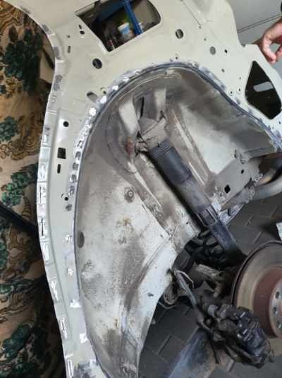 Fragment nadkola samochodu w trakcie naprawy blacharskiej, widoczne ślady spawania i szlifowania, amortyzator i tarcza hamulcowa w tle.