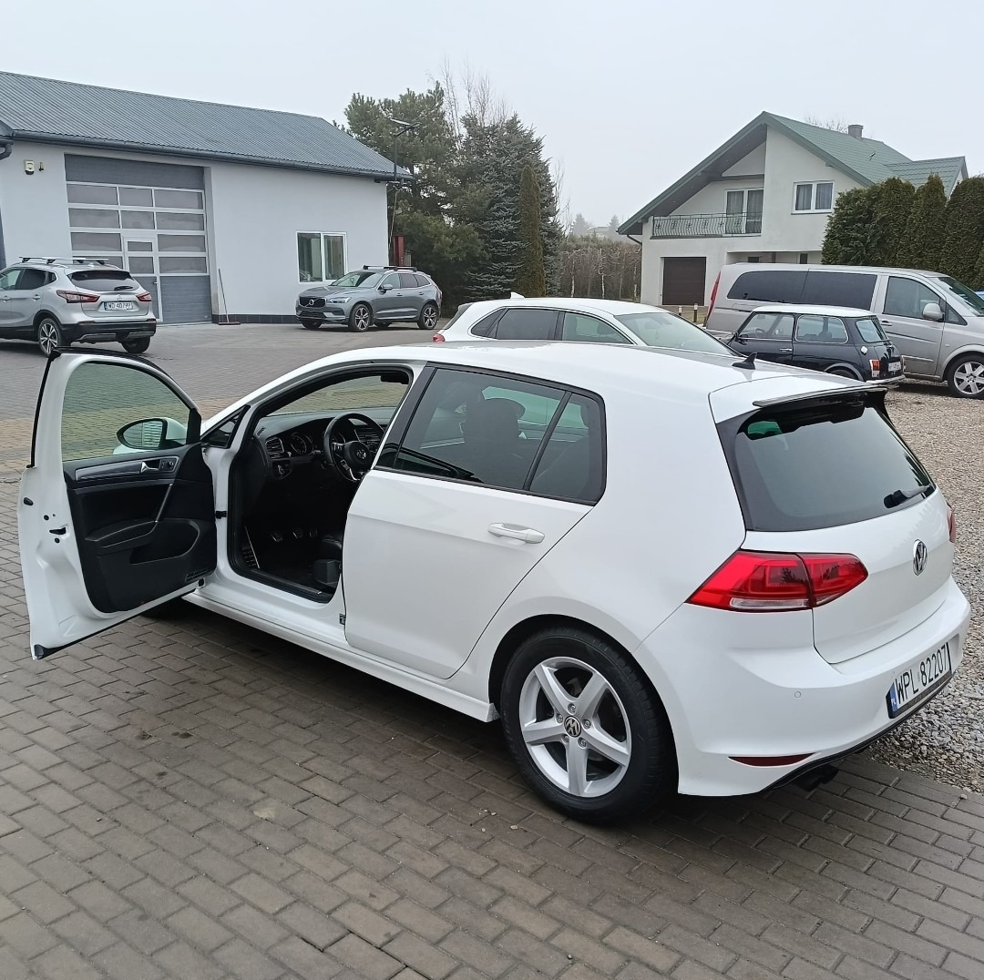 Biały Volkswagen Golf z otwartymi drzwiami kierowcy na brukowanym placu, inne samochody i budynki w tle.