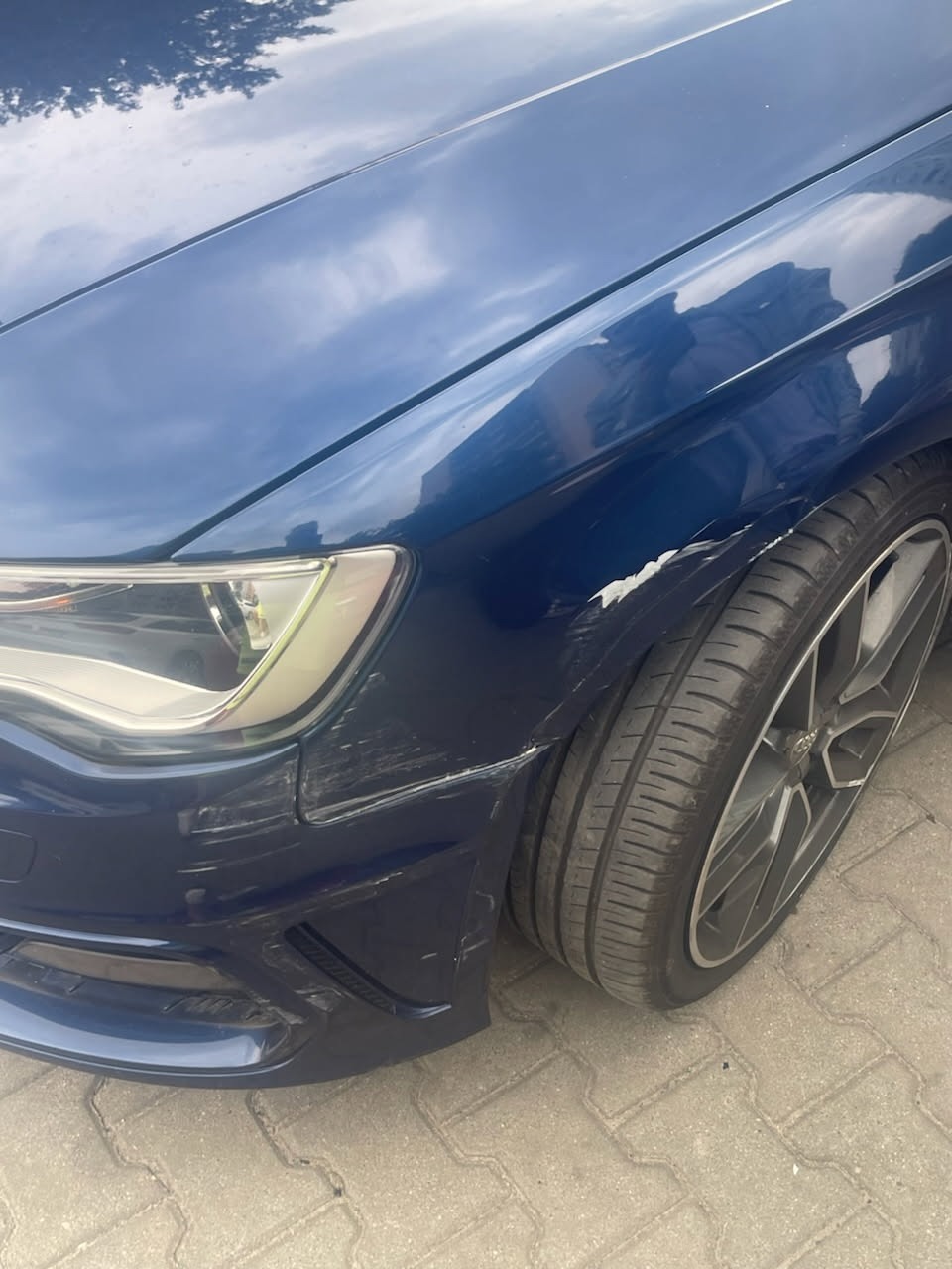 Uszkodzony, niebieski błotnik i zarysowany zderzak samochodu marki Audi, widoczne ślady otarć i odprysków lakieru. Koło z felgą aluminiową.